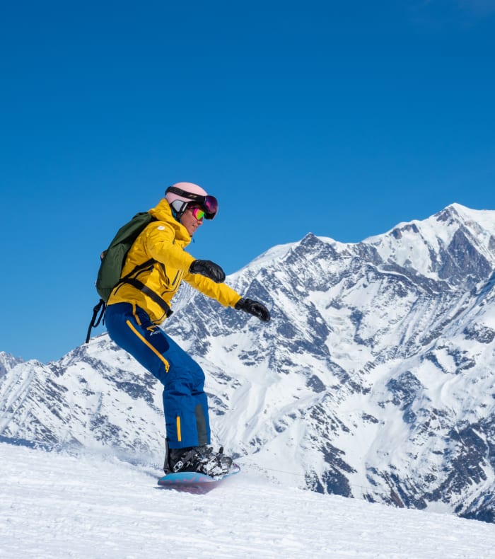 Snowboardeur en veste jaune et pantalon bleu dévale une pente enneigée face aux pics montagneux de Serre Chevalier