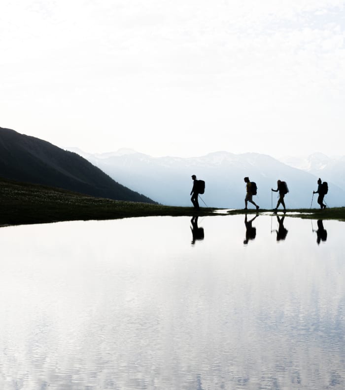 Quatre randonneurs en silhouette traversant un lac de montagne avec sommets enneigés en arrière-plan