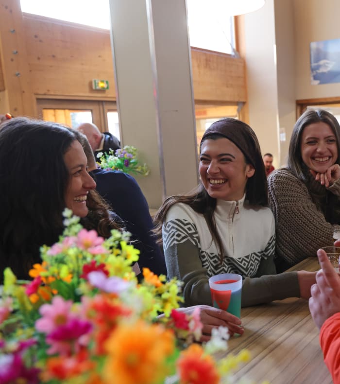 Groupe de skieurs souriants réunis autour d'une table dans un lodge alpin, discutant et partageant des moments de convivialité après une journée hors-piste