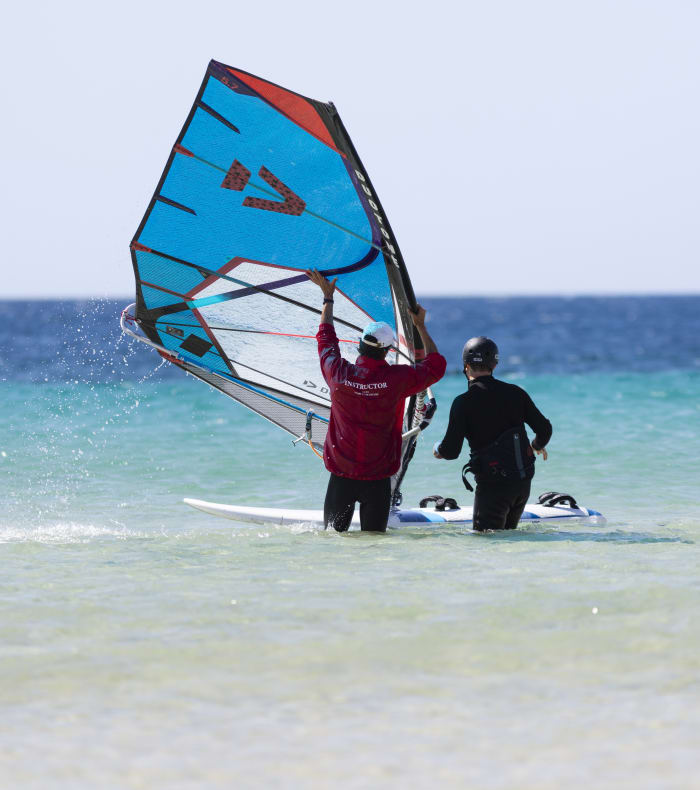Deux pratiquants de windsurf en mer avec voile bleue et rouge, préparant leur sortie en eau turquoise et peu profonde