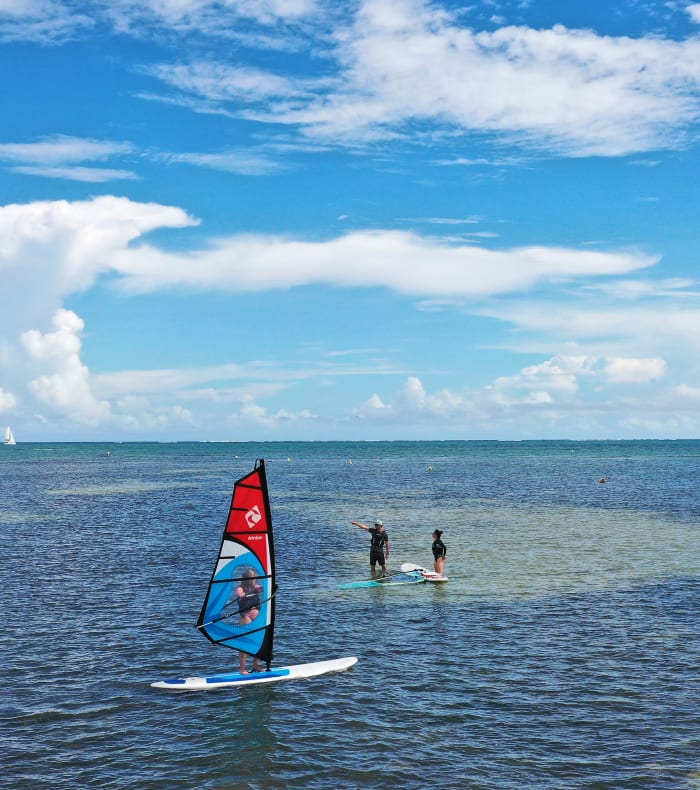 Véliplanchiste en eau turquoise avec voile rouge face à des windsurfeurs en apprentissage sous ciel bleu ensoleillé en Martinique