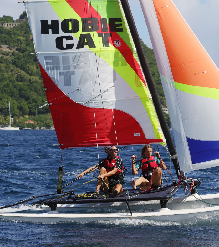 Catamaran blanc avec voiles colorées naviguant en baie des Saintes, deux navigateurs à bord et côte verdoyante en arrière-plan