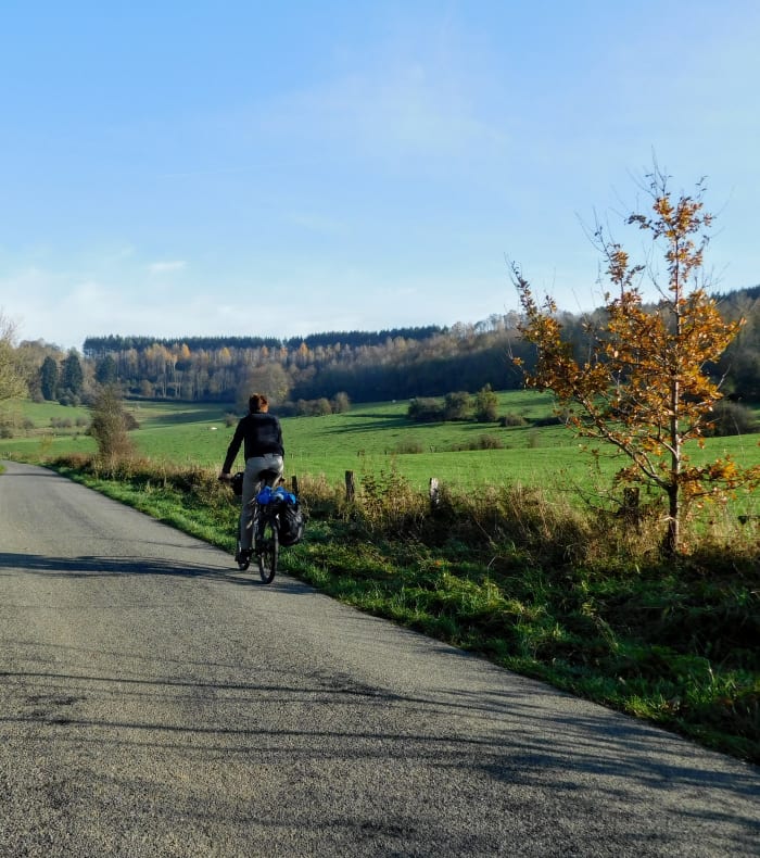 Cycliste en bike packing pédalant sur une route rurale belge avec vue panoramique sur vallées verdoyantes et châteaux