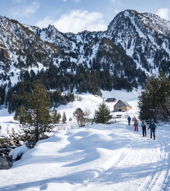 Vallée enneigée des Pyrénées avec randonneurs sur sentier hivernal, maison traditionnelle et pics montagneux boisés au Val d'Aran