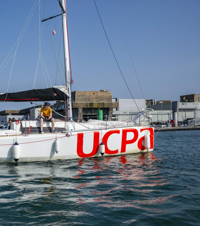 Voilier habitable vue sur le côté du voilier blanc au logo UCPA rouge avec équipage à bord dans la rade de Lorient La Base, stage de perfectionnement en voile niveau 2 FFV