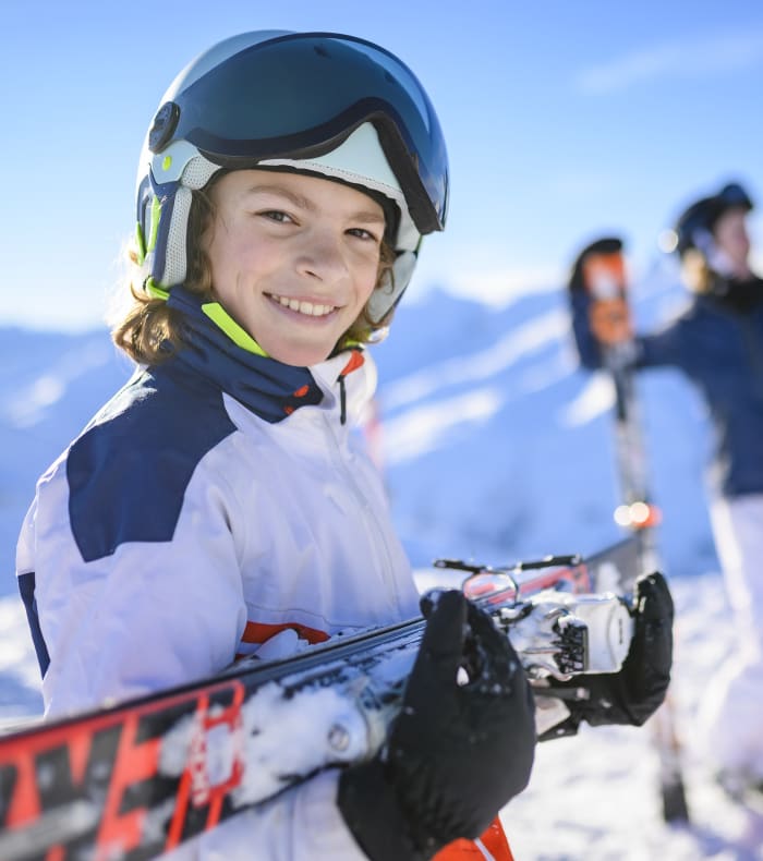 Enfant souriant en tenue de ski avec casque et lunettes tenant ses skis, famille en arrière-plan sur piste enneigée en montagne