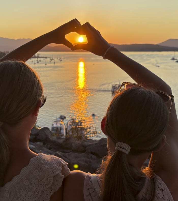 Deux petites filles faisant un cœur avec leur mains où l'on voit apparaitre au centre le soleil au moment du coucher du soleil lors d'une croisière aux Cyclades mer bleue en arrière-plan