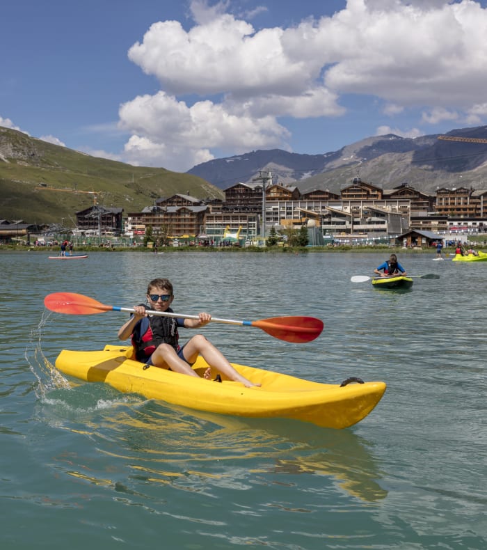 Enfant en kayak jaune sur le lac alpin de Tignes avec montagnes enneigées et station en arrière-plan