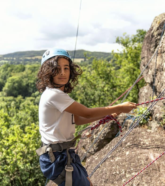 Jeune adolescent en escalade sur paroi rocheuse avec équipement de sécurité et paysage de Normandie en arrière-plan