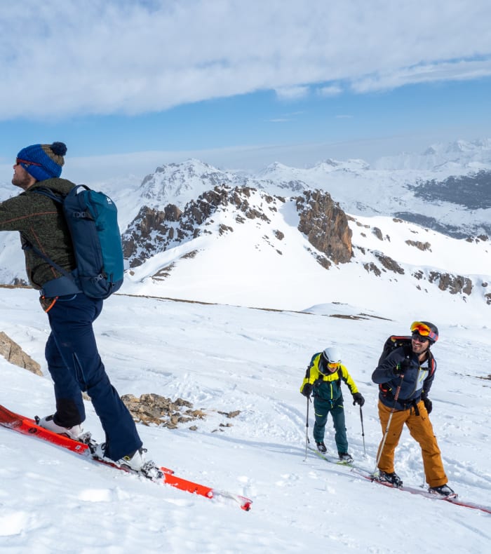 Skieur de randonnée en tenue jaune et bleue montant une pente enneigée avec skis et bâtons de rando en montagne