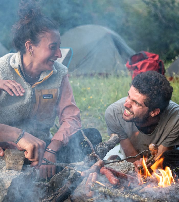 Groupe de trois personnes riant autour d'un feu de camp en montagne, illustrant le bien-être par l'activité sportive et l'engagement de l'UCPA
