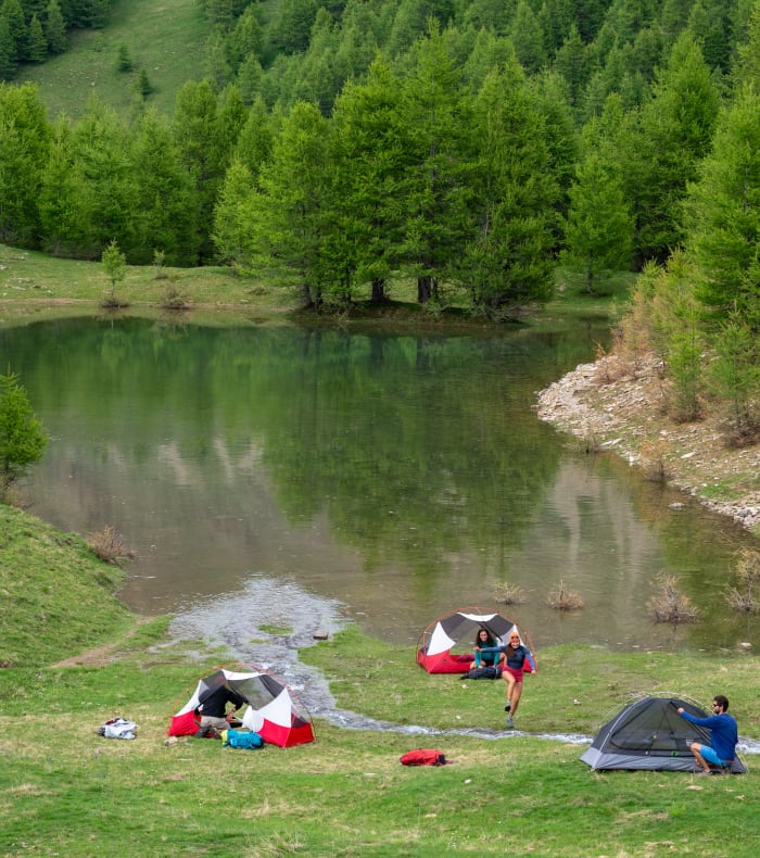 Bivouac en montagne avec tentes rouges et bleues au bord d'un lac alpin entouré de forêts de mélèzes