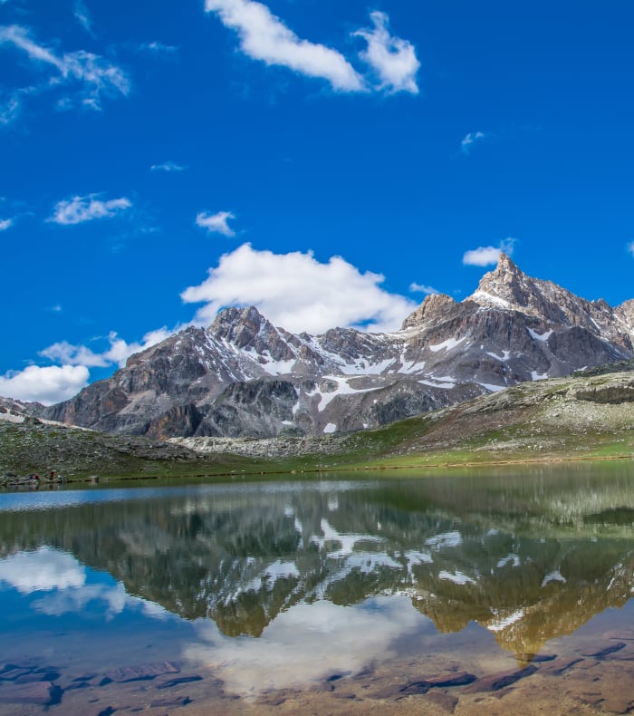 Lac alpin aux Écrins avec pics rocheux enneigés, prairies verdoyantes et eau turquoise sous ciel bleu