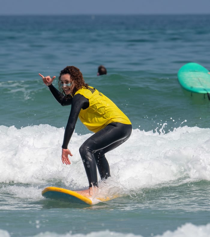 Surfeur en combinaison jaune et noire chevauchant une vague à la Torche, Finistère, avec groupe d'apprentis en arrière-plan