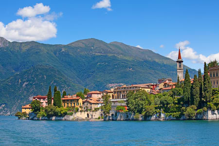Vue panoramique sur le village   pittoresque de Varenna situé au bord du lac de Côme en Italie, avec des bâtiments colorés et une église à clocher pointu, entouré de montagnes verdoyantes sous un ciel bleu lumineux.