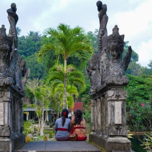 Deux femmes en habits traditionnels assises face à une entrée sculptée d'un temple, entourée de végétation tropicale avec des palmiers et des fleurs colorées à Bali