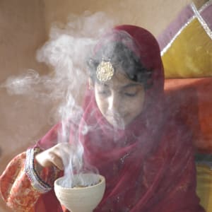 Jeune fille omanaise en tenue traditionnelle rouge, manipulant un encensoir en céramique d'où s'élèvent des volutes de fumée parfumée