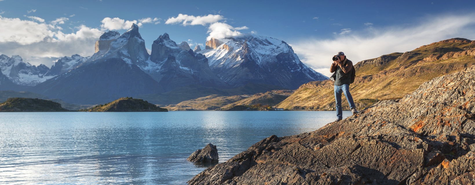Un photographe prend des photos posé au bord d'un lac, avec des montagnes enneigées en arrière plan, dans le Parc national de Torres del Paine, Patagonie