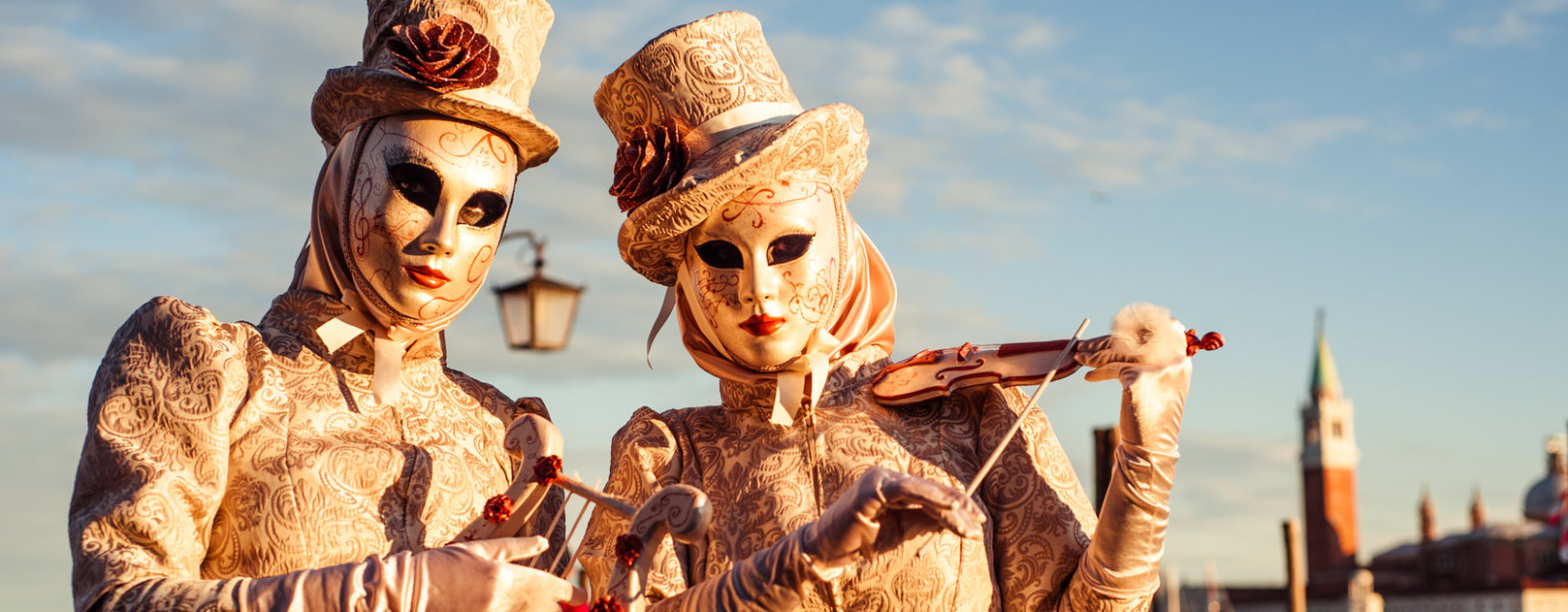 Deux participants du Carnaval de Venise, habillés en costumes traditionnels brodés d'or avec des masques, posent dans la lumière dorée de fin d'après-midi. L'un tient un violon miniature, ajoutant une touche artistique à l'ensemble.