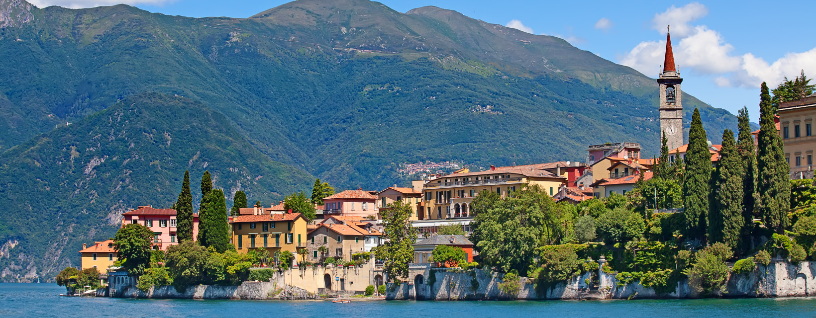 Vue panoramique sur le village   pittoresque de Varenna situé au bord du lac de Côme en Italie, avec des bâtiments colorés et une église à clocher pointu, entouré de montagnes verdoyantes sous un ciel bleu lumineux.