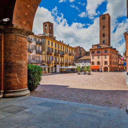View of cobblestone town square among old houses and medieval towers under beautiful sky in Alba, Piedmont, Northern Italy.