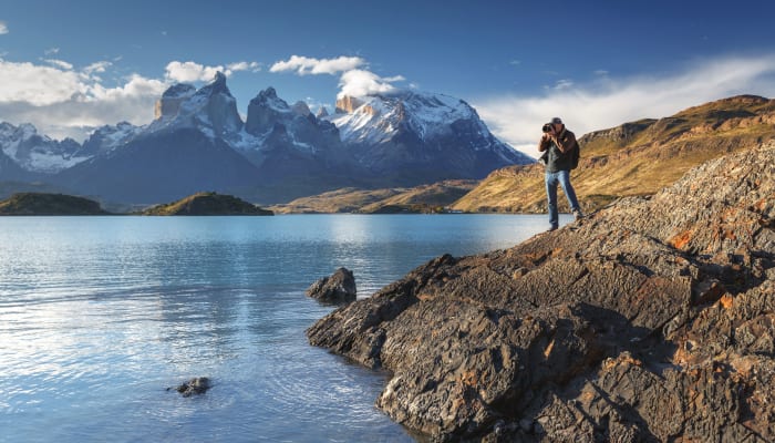Un photographe prend des photos posé au bord d'un lac, avec des montagnes enneigées en arrière plan, dans le Parc national de Torres del Paine, Patagonie